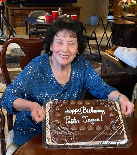 Pastor Joyce smiling behind a chocolate birthday cake that reads Happy Birthday Pastor Joyce